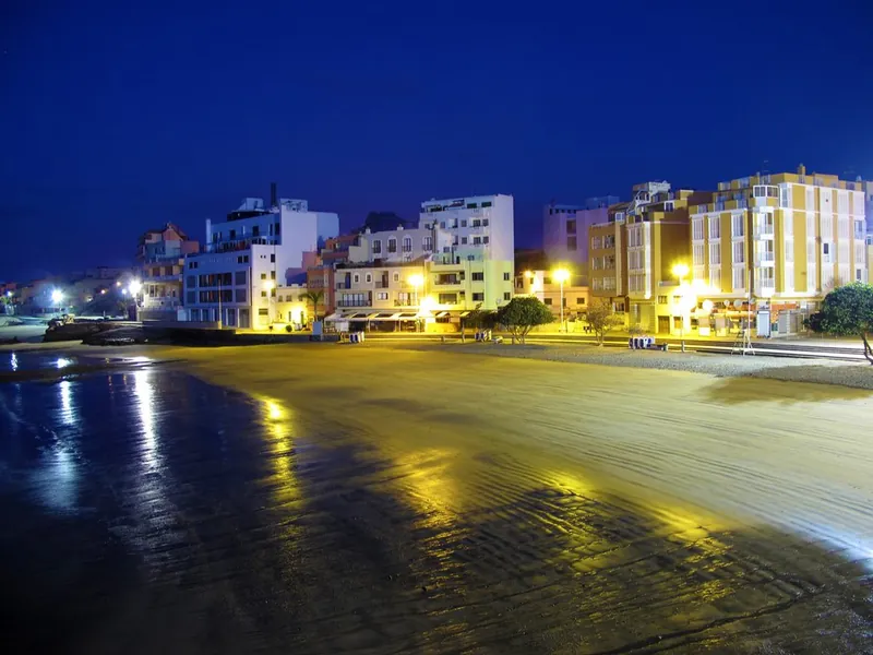 The illuminated seafront of El Médano in the evening, with lights reflected on the wet sand