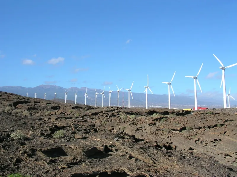 A long row of wind turbines on a dark lava field at the Granadilla Wind Farm