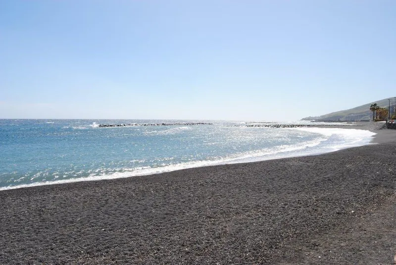 The long black-sand shore of Playa del Cabezo, with the sparkling sea and the breakwater