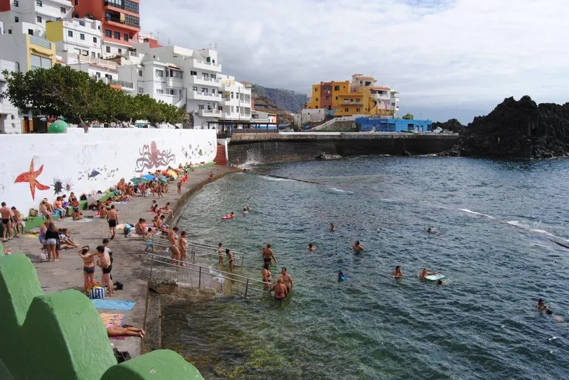Bathers in the natural pool at Playa El Pris, among colourful houses and a sea-themed mural