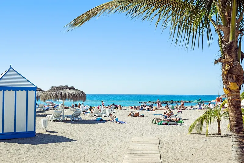 A blue-striped beach hut, a straw parasol and bathers on the sand at Playa de Fañabé