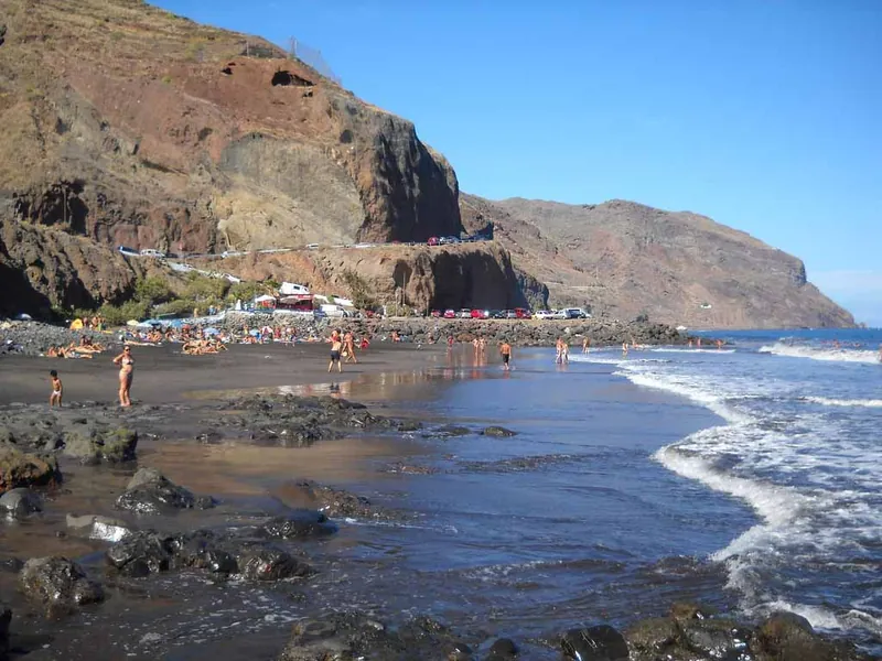 Bathers on the black sand of Playa Las Gaviotas below the cliffs, with parked cars