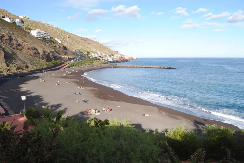 Black sand and a breakwater at Playa de la Nea, with hillside houses and the ocean
