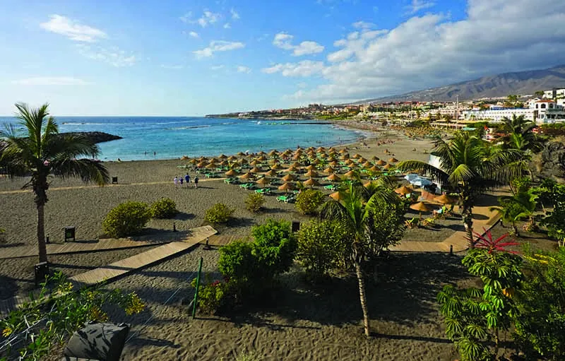 Straw parasols on Playa de Troya seen from the promenade through palms and shrubs